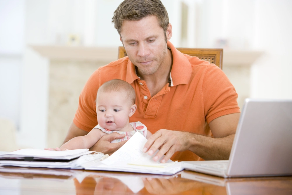 Father sat with baby at a table looking at paperwork with the laptop open