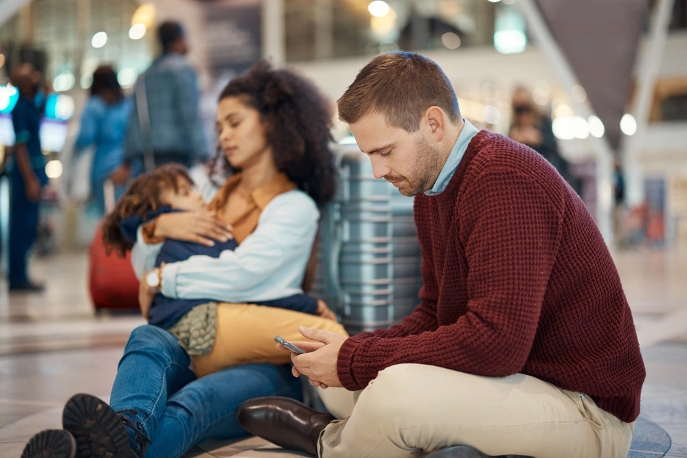Family of three sat on the floor at the airport with the father looking at his phone.