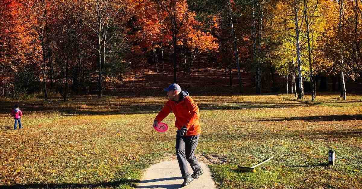 A disc golfer on a tee pad in the open in the middle of a throw that will go into a wooded area ahead of him