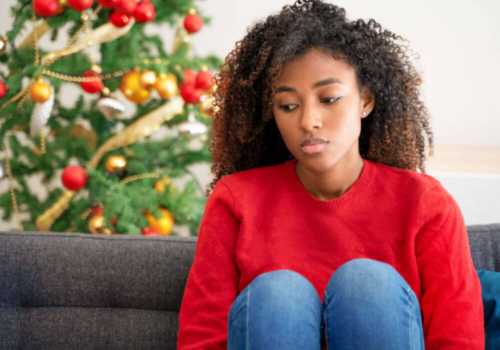 A young black woman sits near a Christmas tree and struggles with a holiday financial challenge.