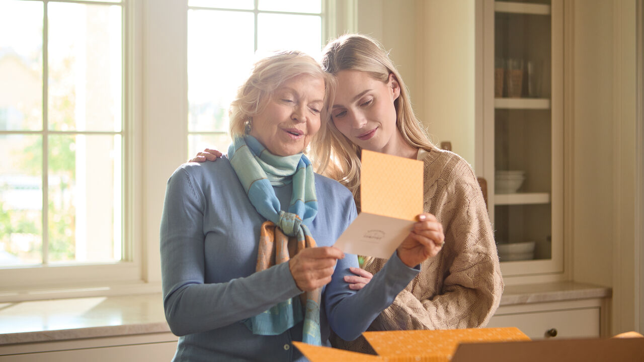 Two women standing close together in a bright kitchen, smiling as they read a greeting card from an open gift box.