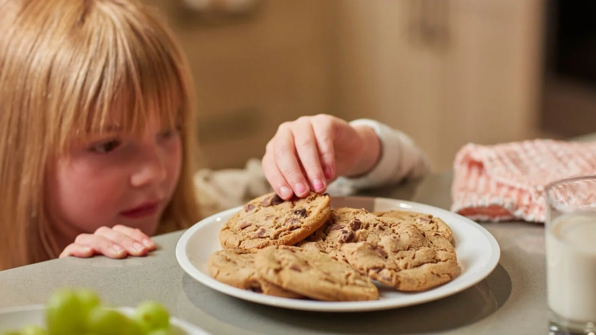 Young child reaching for a chocolate chip cookie from a plate on the table, with a glass of milk nearby.