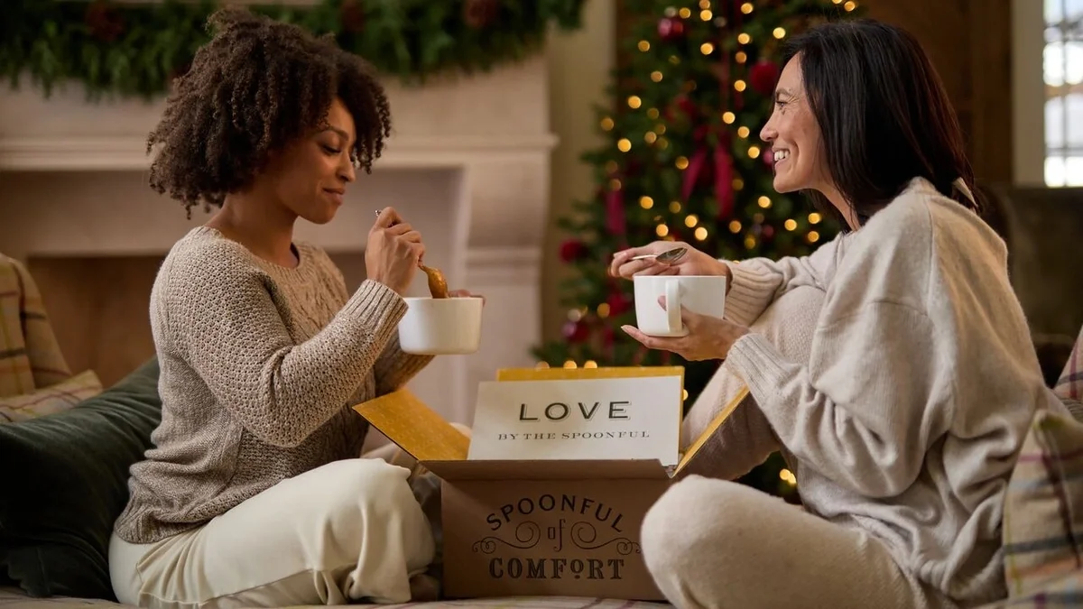 Two women sitting together near a decorated Christmas tree, enjoying warm soup and cookies beside an open Spoonful of Comfort gift box with a “Love by the Spoonful” card.