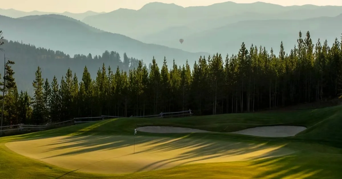 The green of a golf holes with mountains in the distance and a hot air balloon as a tiny speck