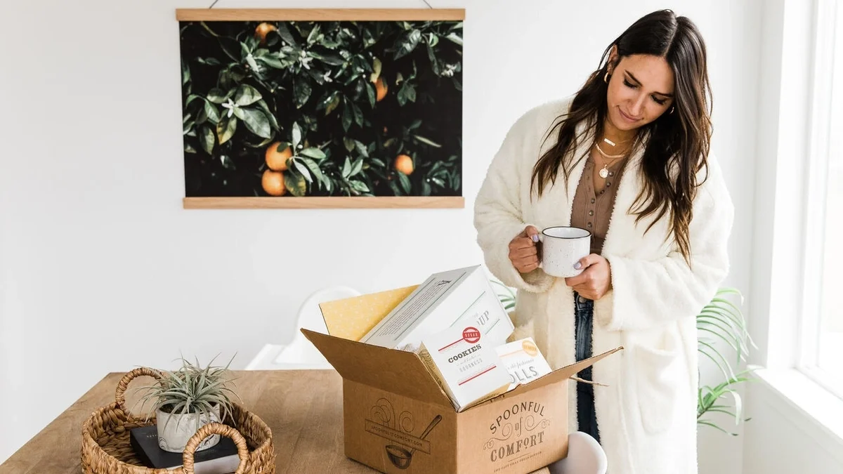 A woman in a cozy white cardigan holds a mug beside an open Spoonful of Comfort gift box filled with packaged treats and gifts.