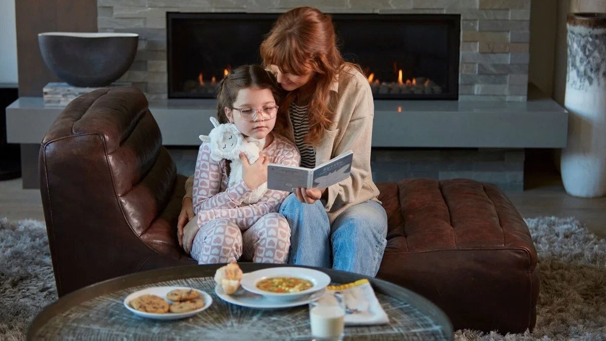 Adult and child sitting together in front of a fireplace, reading a book while cuddling a plush toy, with a bowl of soup, cookies, and milk on the coffee table nearby.