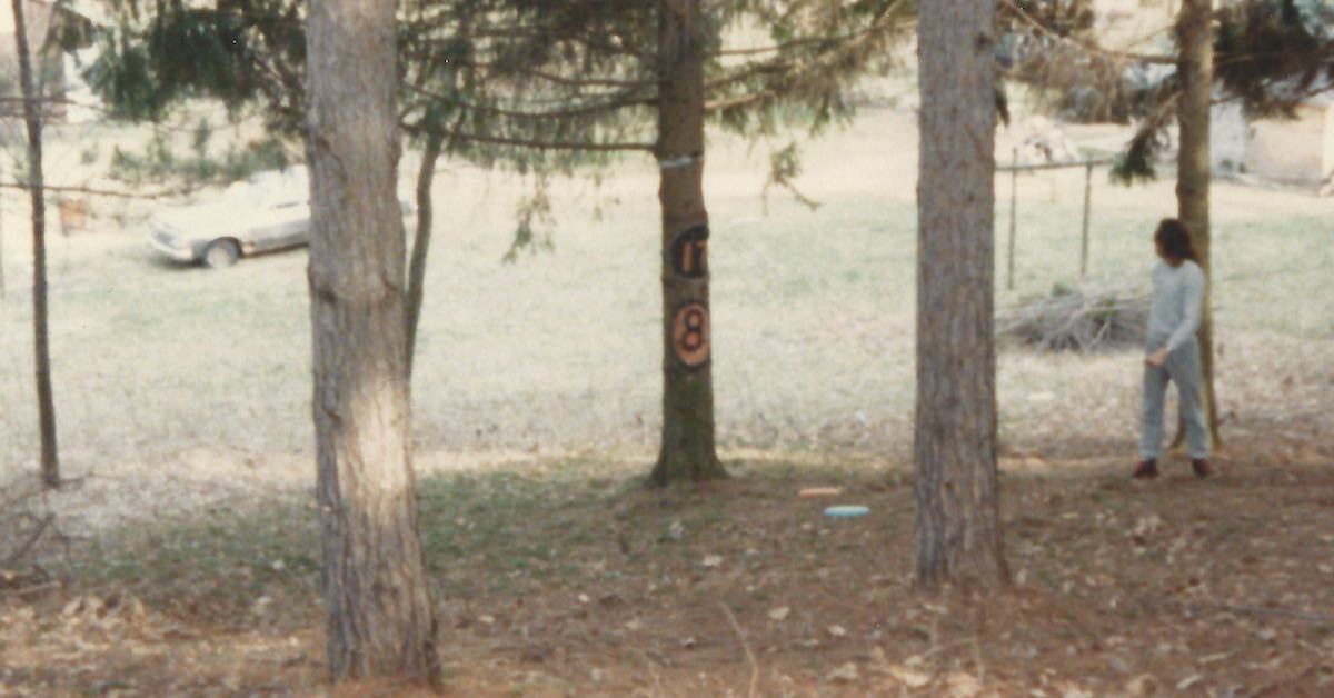 An older photograph of a man in 80s attire playing disc golf in a small patch of trees with an older car in the background
