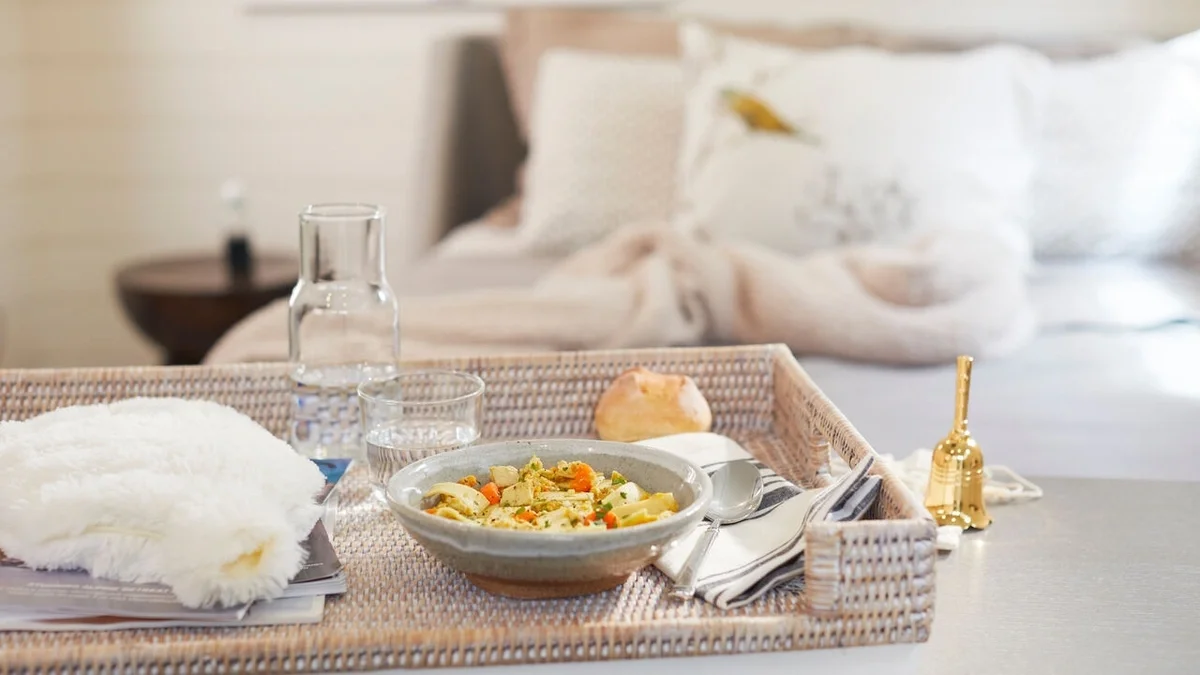 Tray arranged with a bowl of chicken soup, a roll, a glass carafe of water, silverware, a cozy throw blanket, and a gold bedside bell, with a neatly made bed in the background.