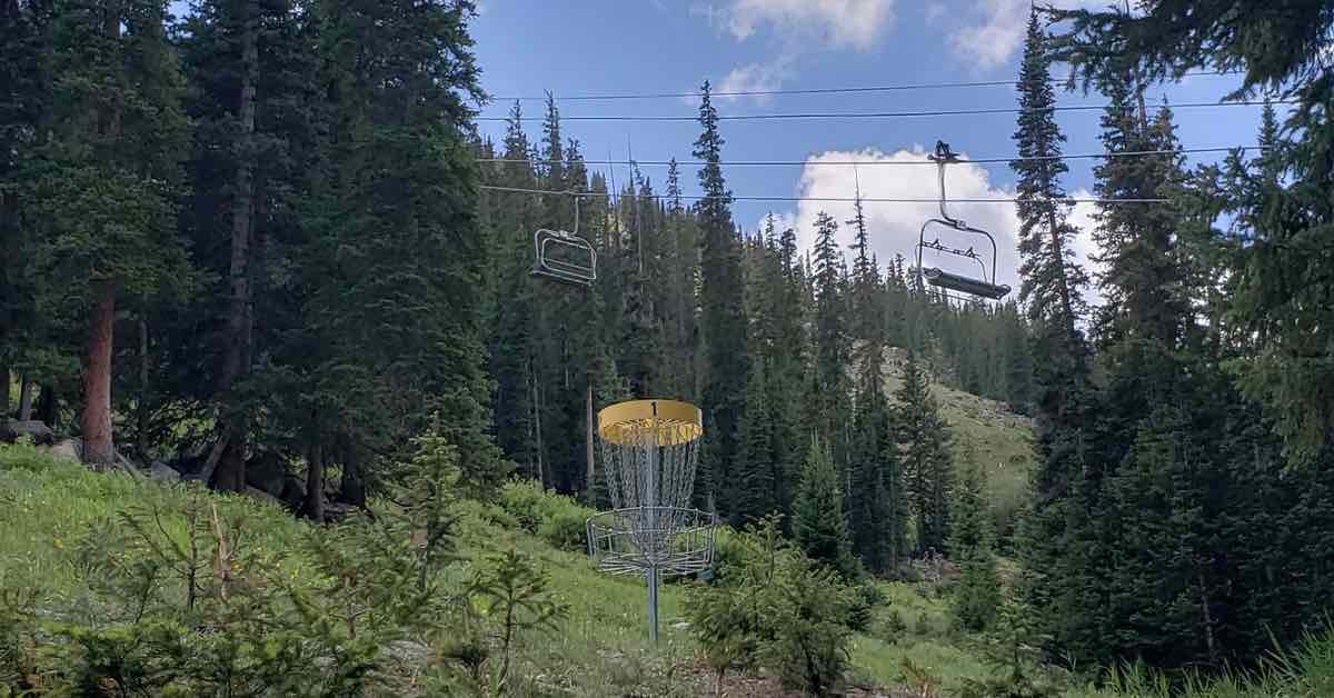 A disc golf basket with a yellow band in a mountain forest. Ski chairlifts hang above it.