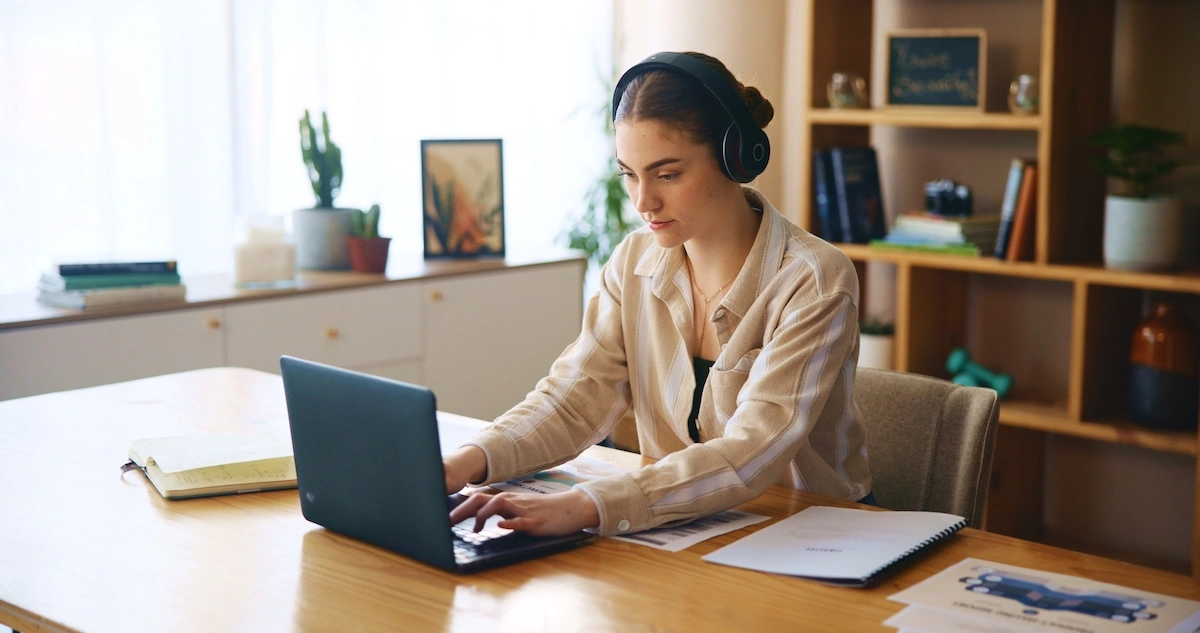 A woman sits in front of a laptop wearing headphones and typing.