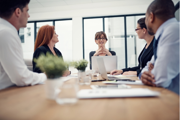 Five office workers sit around a conference room table, the woman in the middle holds hands together in thought while speaking to the group, there is an open computer in front of her