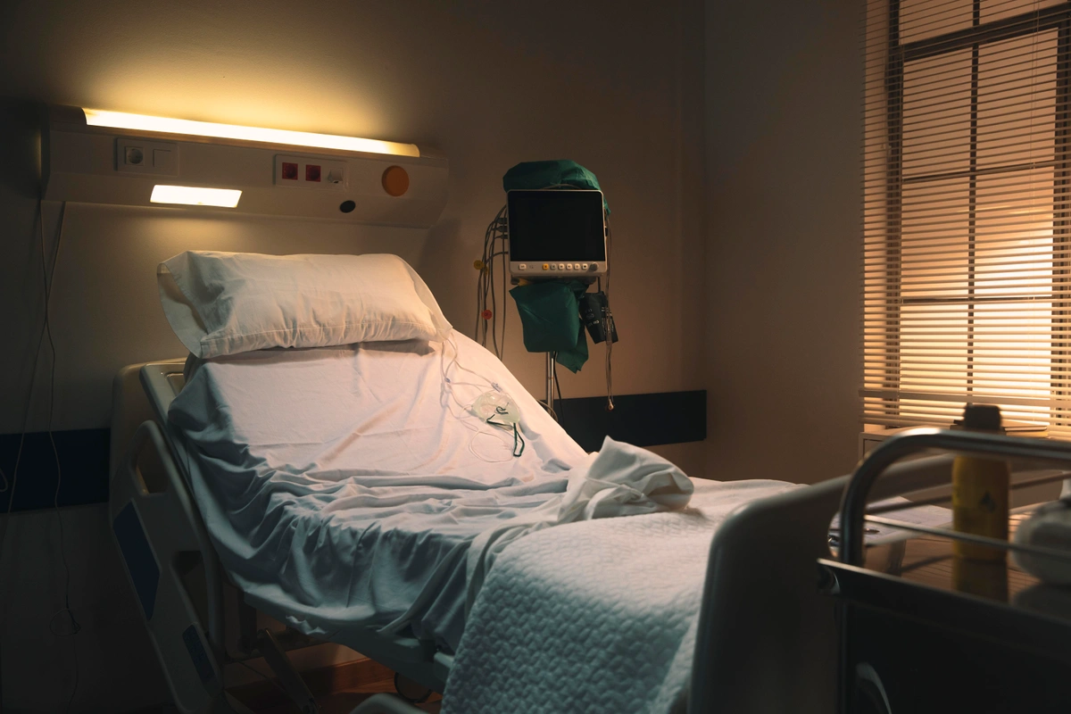 An empty hospital bed in a dimly lit room, with medical equipment and a monitor beside it, giving a somber and quiet atmosphere.