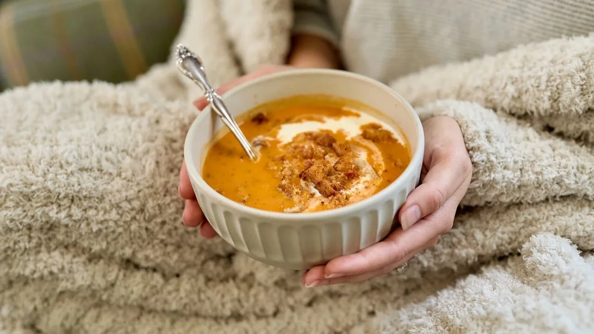 Close-up of hands holding a bowl of creamy tomato soup topped with a swirl of cream and crunchy garnish while wrapped in a cozy throw blanket.