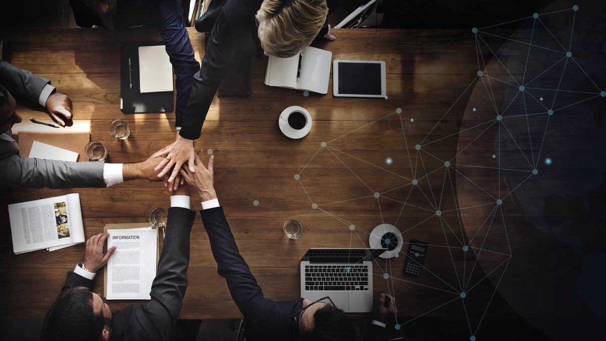Aerial view of a business team around a wooden table, putting their hands together in a gesture of unity, with documents, coffee, and electronic devices on the table.
