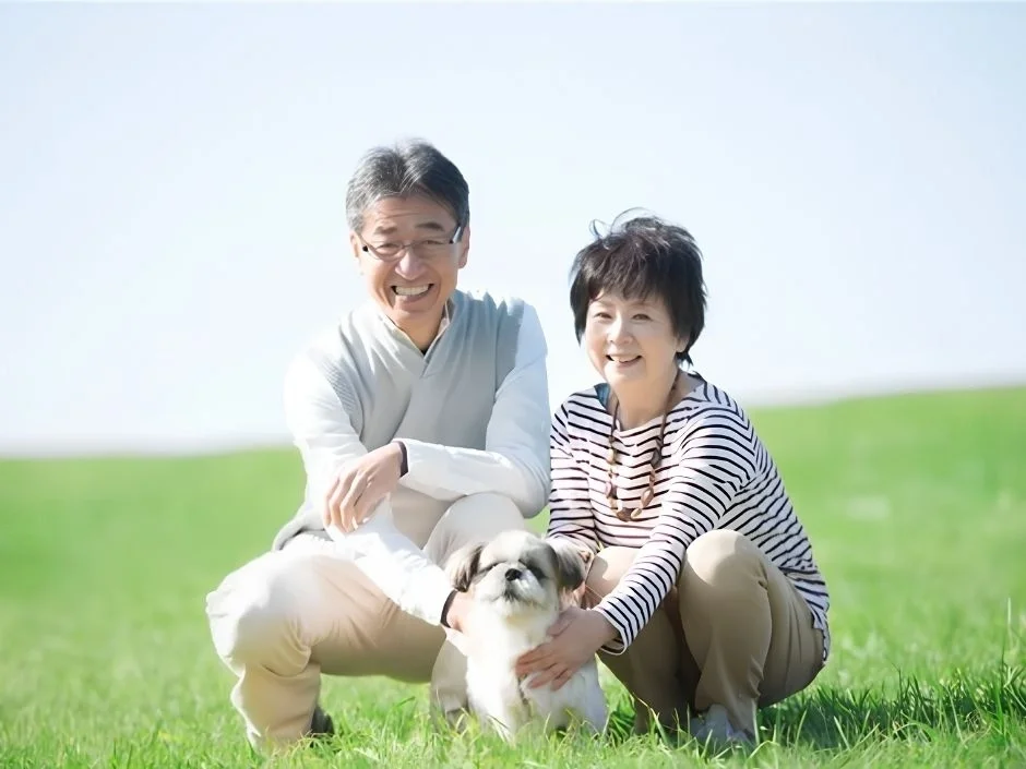 Older Asian couple smiling and kneeling with a small Shih Tzu puppy in a bright green field