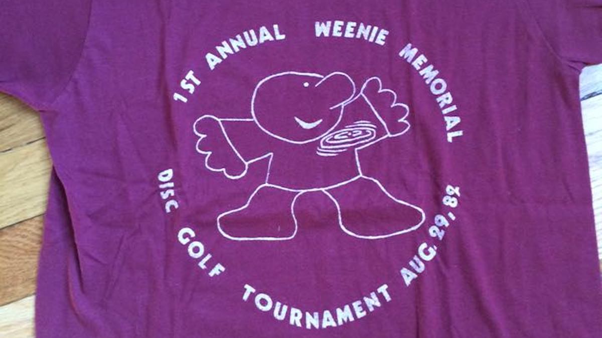 Purple shirt with rudimentary sketch of a cartoon man playing disc golf in white. Shirt says "Weenie Memorial" as weel as other text.