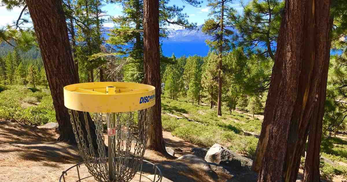 A disc golf basket on a ridge with a view over evergreen trees down to mountains surrounding a large lake