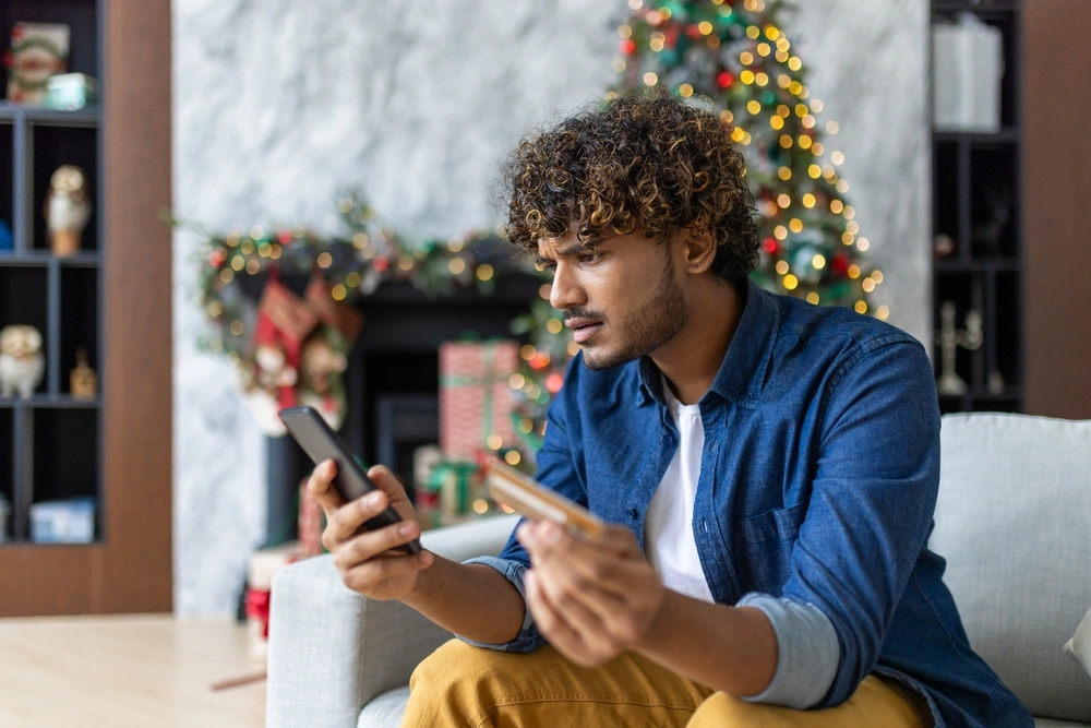 Man sat in living room which is decorated for Christmas. He's holding his phone and credit card and looking concerned.