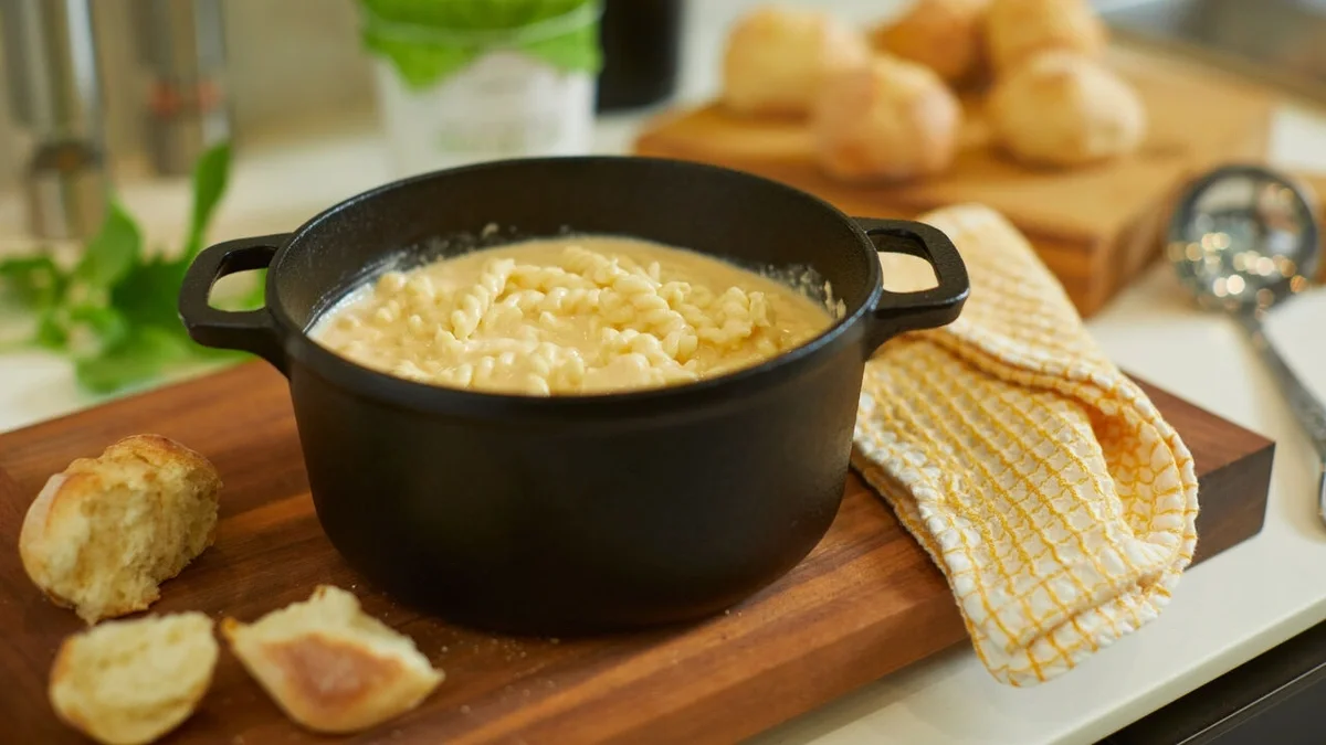Black Dutch oven filled with creamy macaroni and cheese on a wooden board, surrounded by torn rolls, a yellow kitchen towel, and serving utensils.