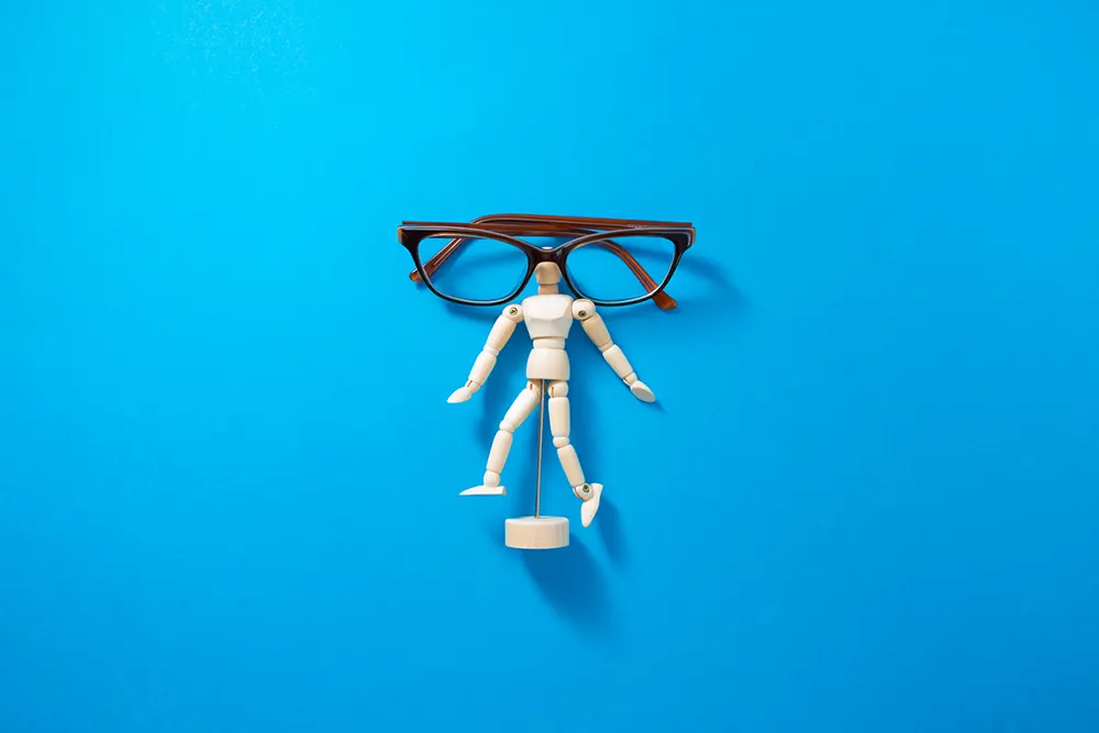 Image shows a miniature posable wooden figure-model, posed arms-open on a plain blue background. Over the head rests a folded women's reading glasses with brown frames. 