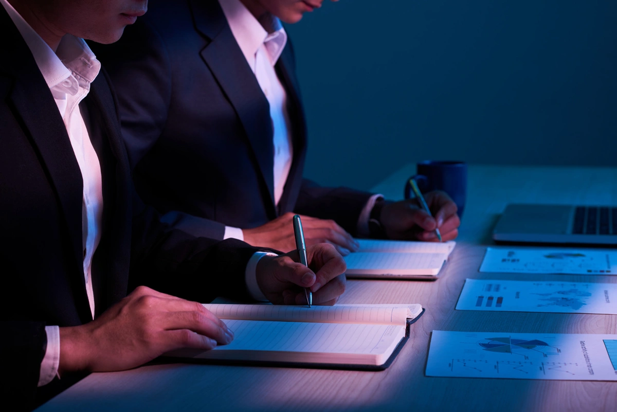  Two individuals in business attire seated side by side in a dimly lit environment, writing notes in notebooks. On the table are charts, graphs, and a laptop, illuminated by soft blue lighting, suggesting a focused work session or analysis.
