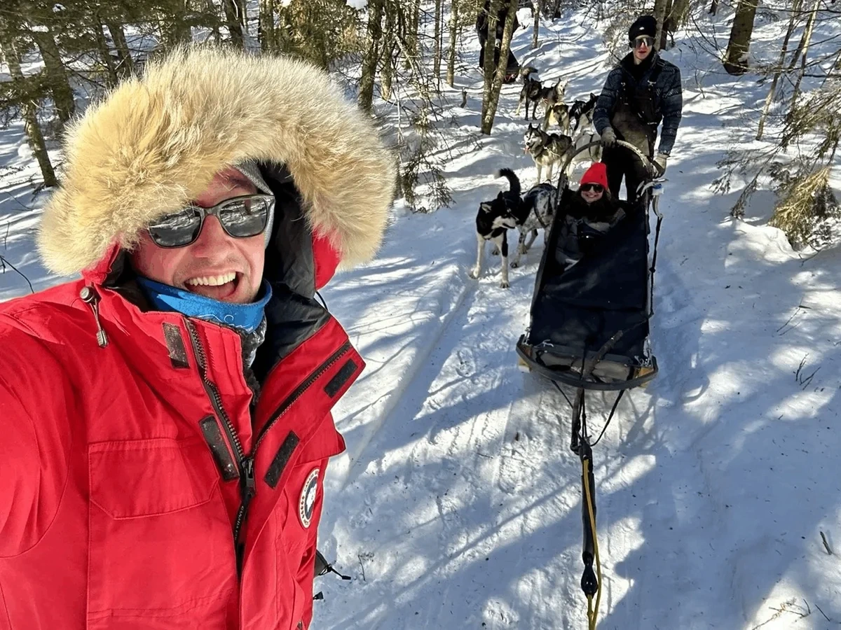 Three people in red and black coats pose with a team of sled dogs in the snow.