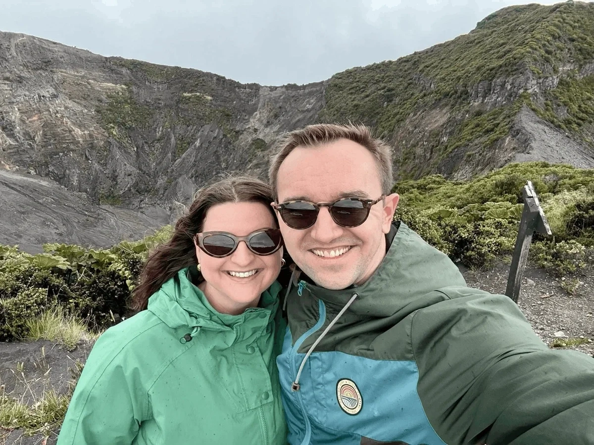 Travel bloggers Christopher and Bri pose in green jackets in front of a volcano crater in Costa Rica.