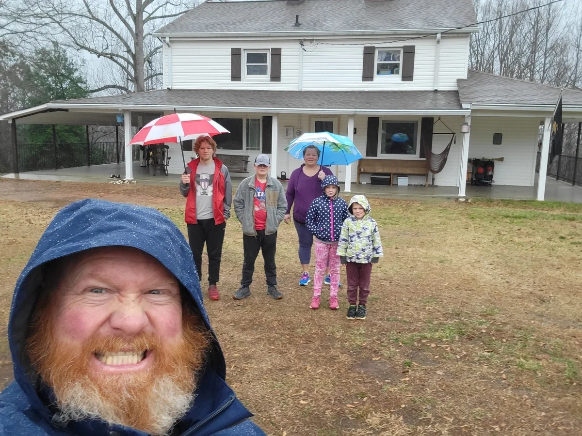 Six members of the Powers family stand in front of a white farmhouse on a rainy day.