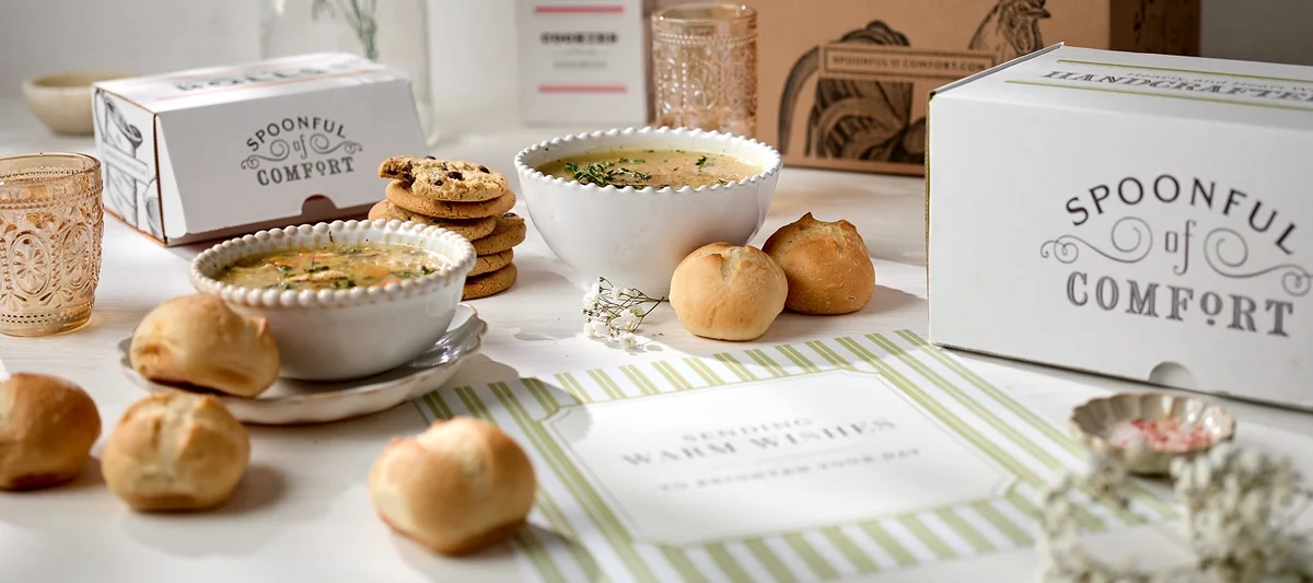 A styled tabletop display with two bowls of soup topped with herbs, small bread rolls, stacked cookies, and branded packaging boxes, arranged alongside a “Happy Mother’s Day” card on a light surface.