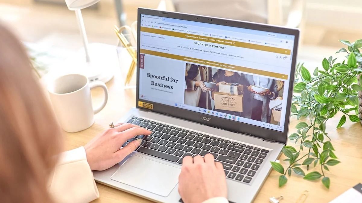 Person browsing the Spoonful of Comfort website on a laptop at a bright desk, with a coffee mug and green plant nearby.