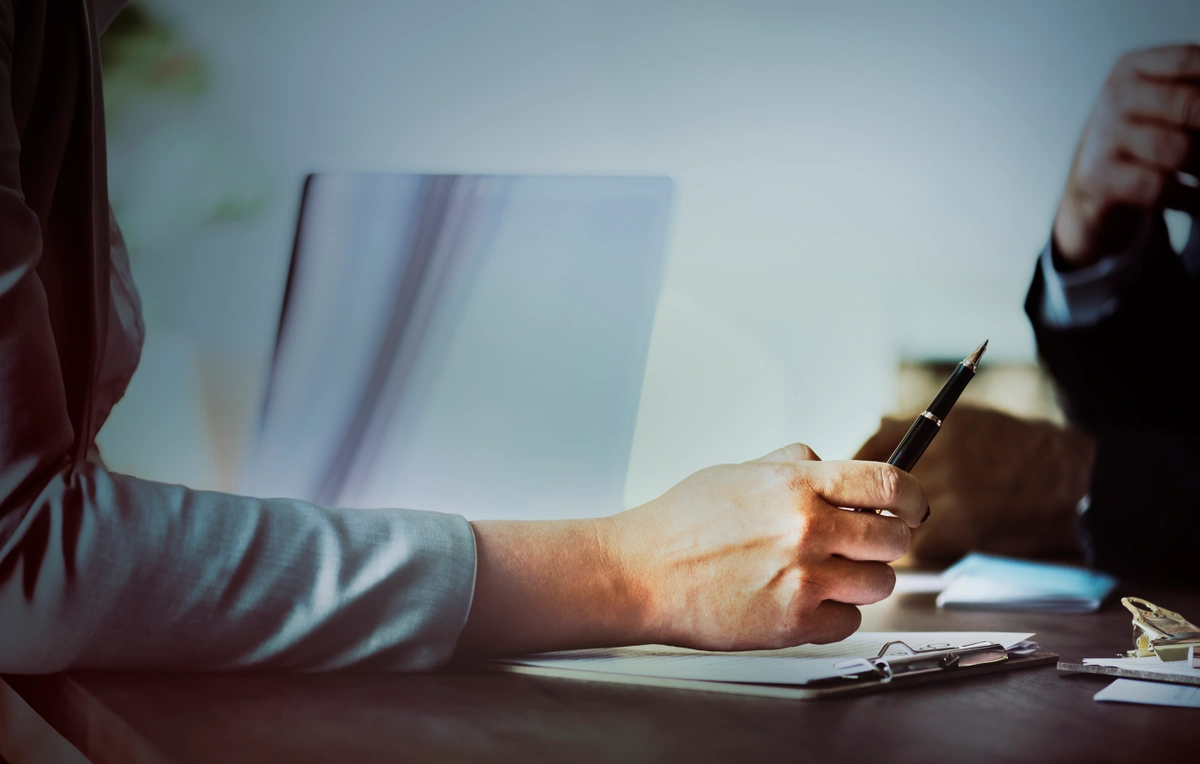 Close-up of a business person holding a pen, with a laptop and documents on the table, during a meeting.