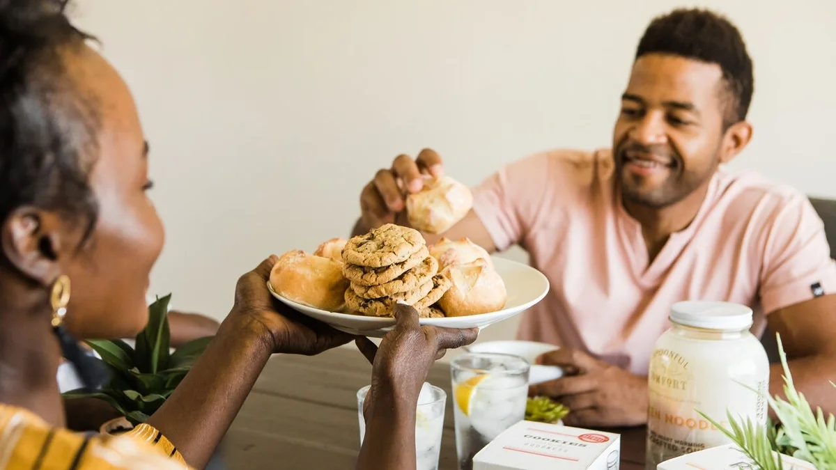 Two people sitting together at a table, smiling as they share a plate piled with rolls and cookies, with Spoonful of Comfort items nearby.