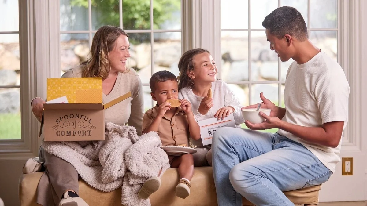 Family sitting together in a bright room, smiling and sharing soup while a child enjoys a roll, with an open Spoonful of Comfort gift box and cozy blanket nearby.