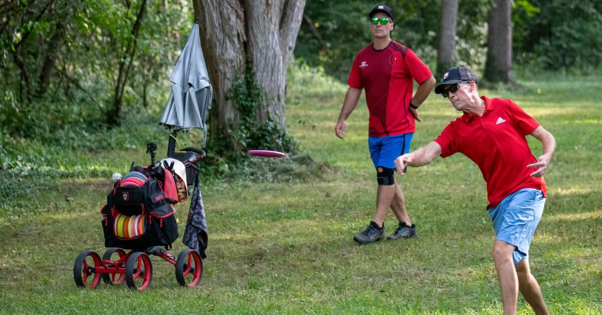 Two older male disc golfers, one throwing, both with red tee shirts on.