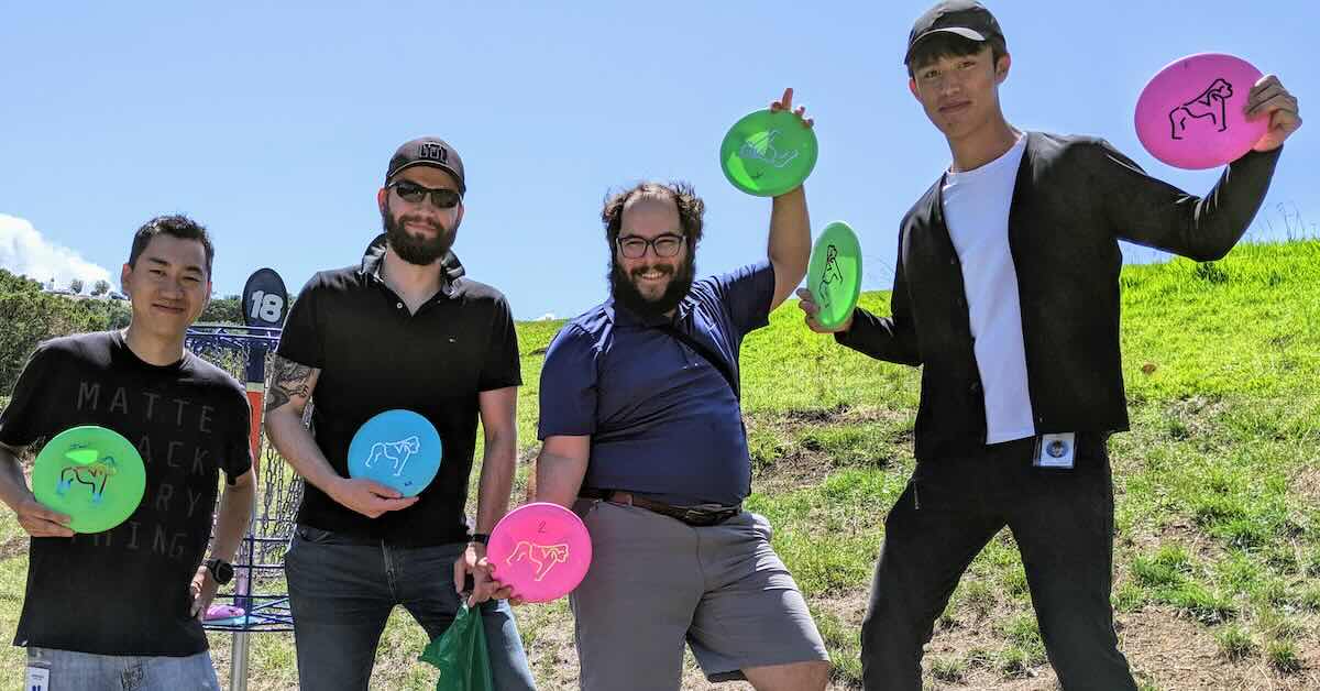 Four men near a disc golf basket holding discs
