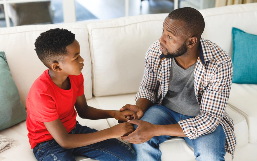 Serious african american father and son sitting on couch in living room talking and holding hands