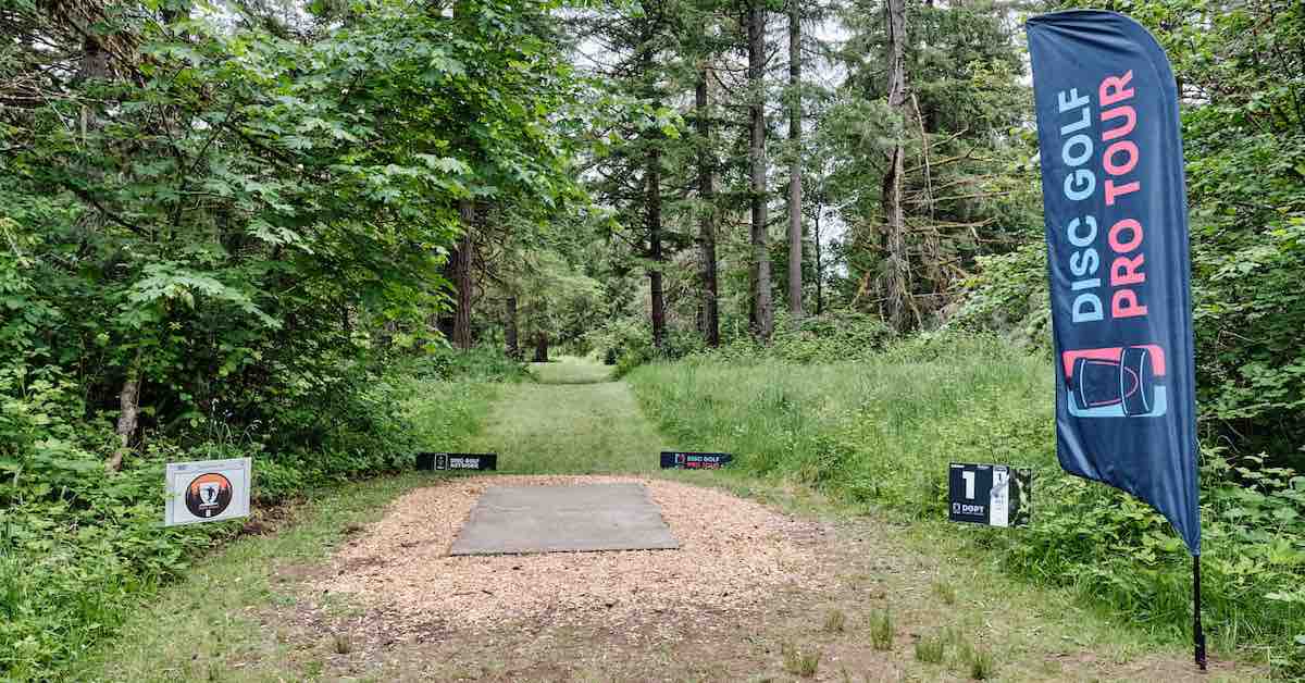 A concrete disc golf tee pad leading to a wooded fairway. A Disc Golf Pro Tour flanner is in the foreground on the right