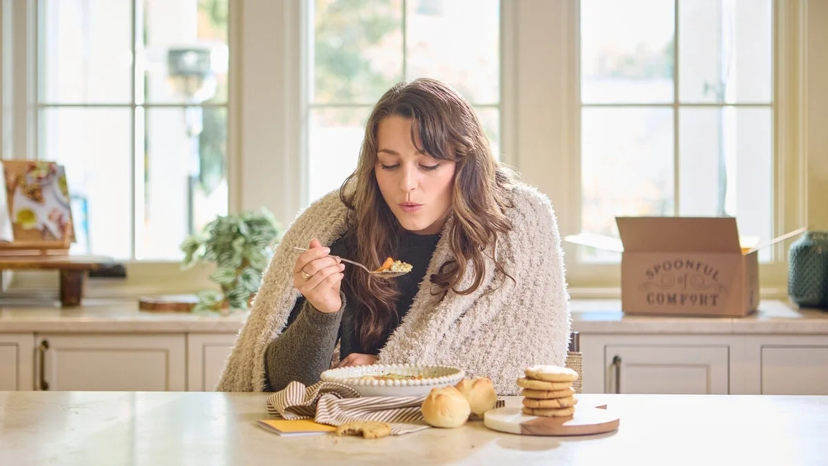 Woman wrapped in a cozy blanket, enjoying a bowl of soup at a kitchen table with rolls and cookies nearby, while an open Spoonful of Comfort gift box sits in the background.