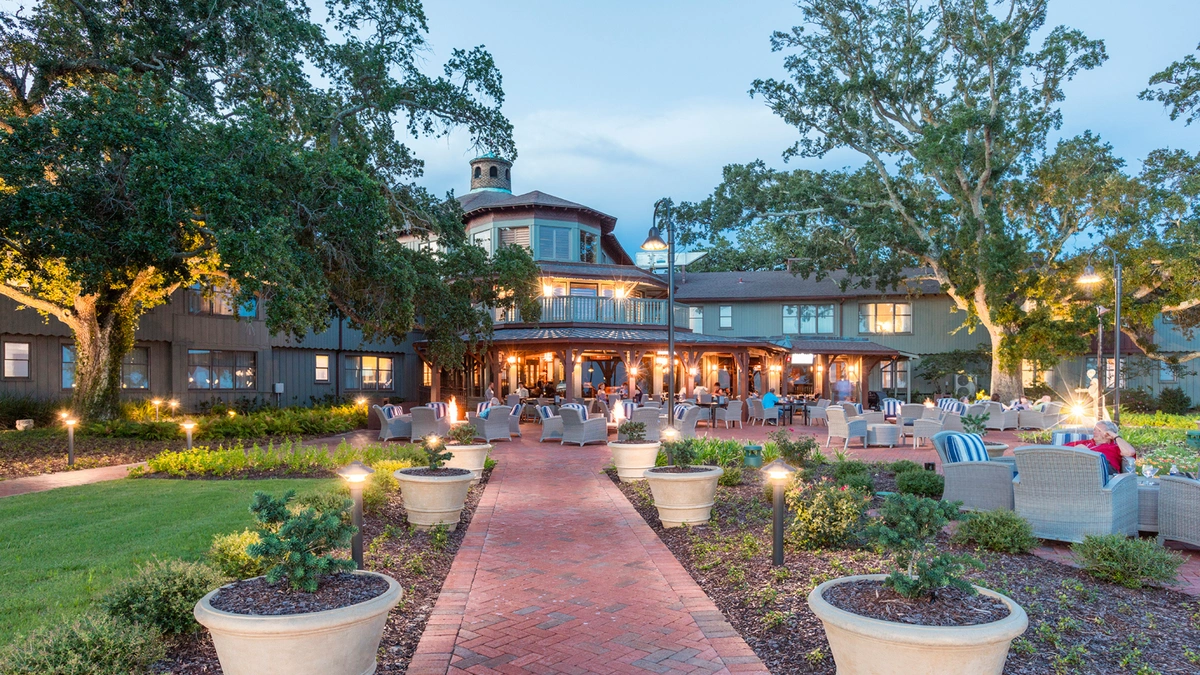 An outdoor patio in front of a large building, surrounded by greenery and tall trees. The patio features numerous seating areas with chairs and tables.