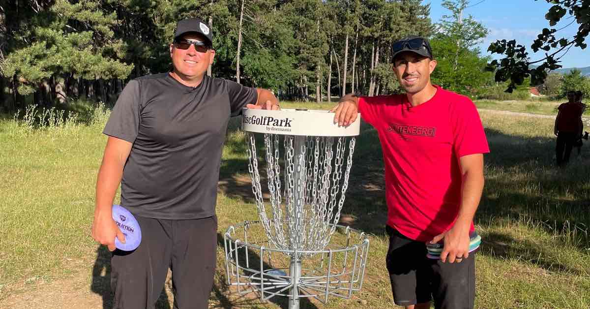 Two men smiling for the camera while leaning on a disc golf basket