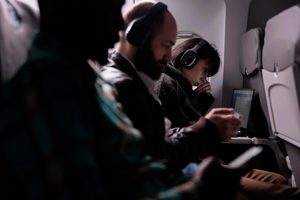 Three airplane passengers wearing headphones; one uses a phone, another focuses on their hands, and the third works on a laptop by the window in dim lighting.