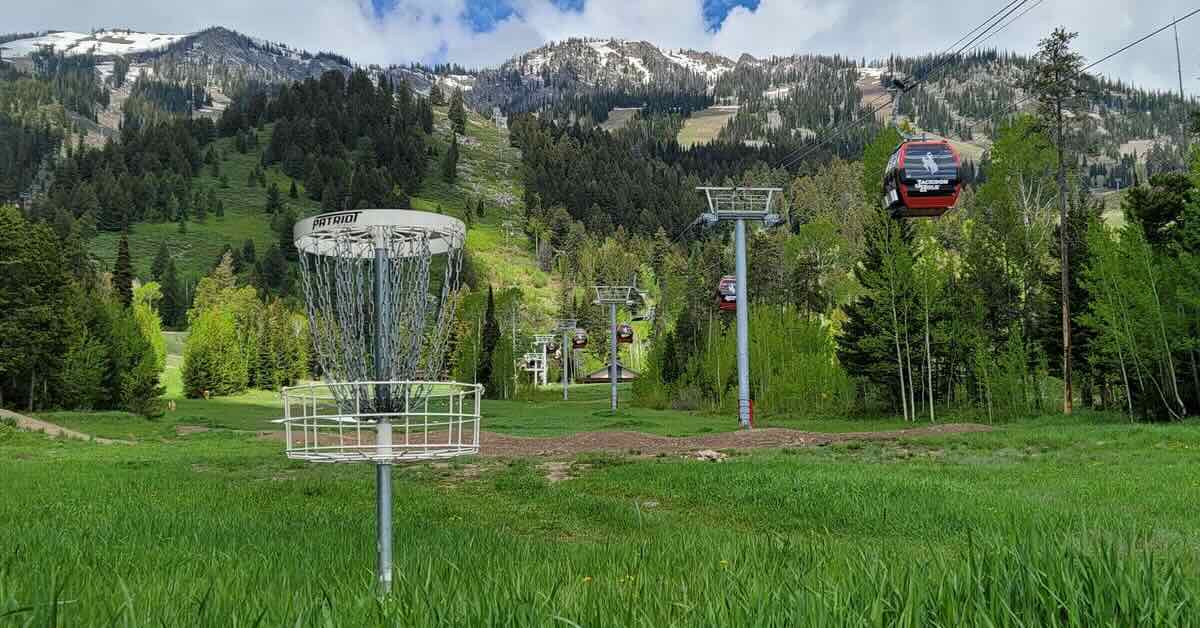 A disc golf basket with ski lifts and green mountains with snowy tops