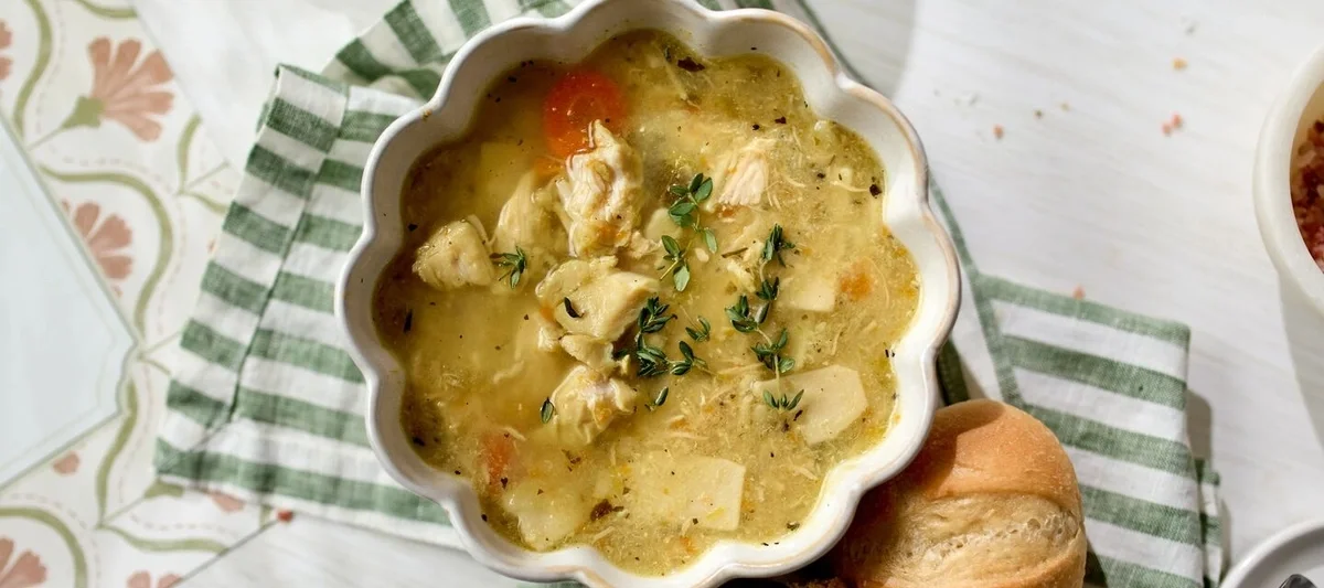 Close-up of a bowl of chicken soup with shredded chicken, carrots, herbs, and broth, set on a green striped cloth with a roll nearby.