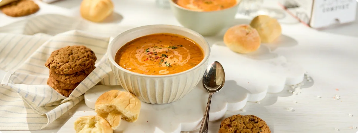 Bowl of creamy tomato soup garnished with herbs, surrounded by bread rolls and cookies on a white table.
