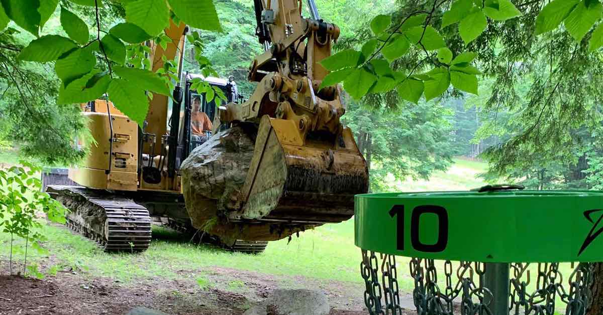 An excavator with a large boulder in its shovel in a wooded area