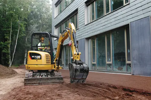 A person operating a mini excavator on dirt near a building