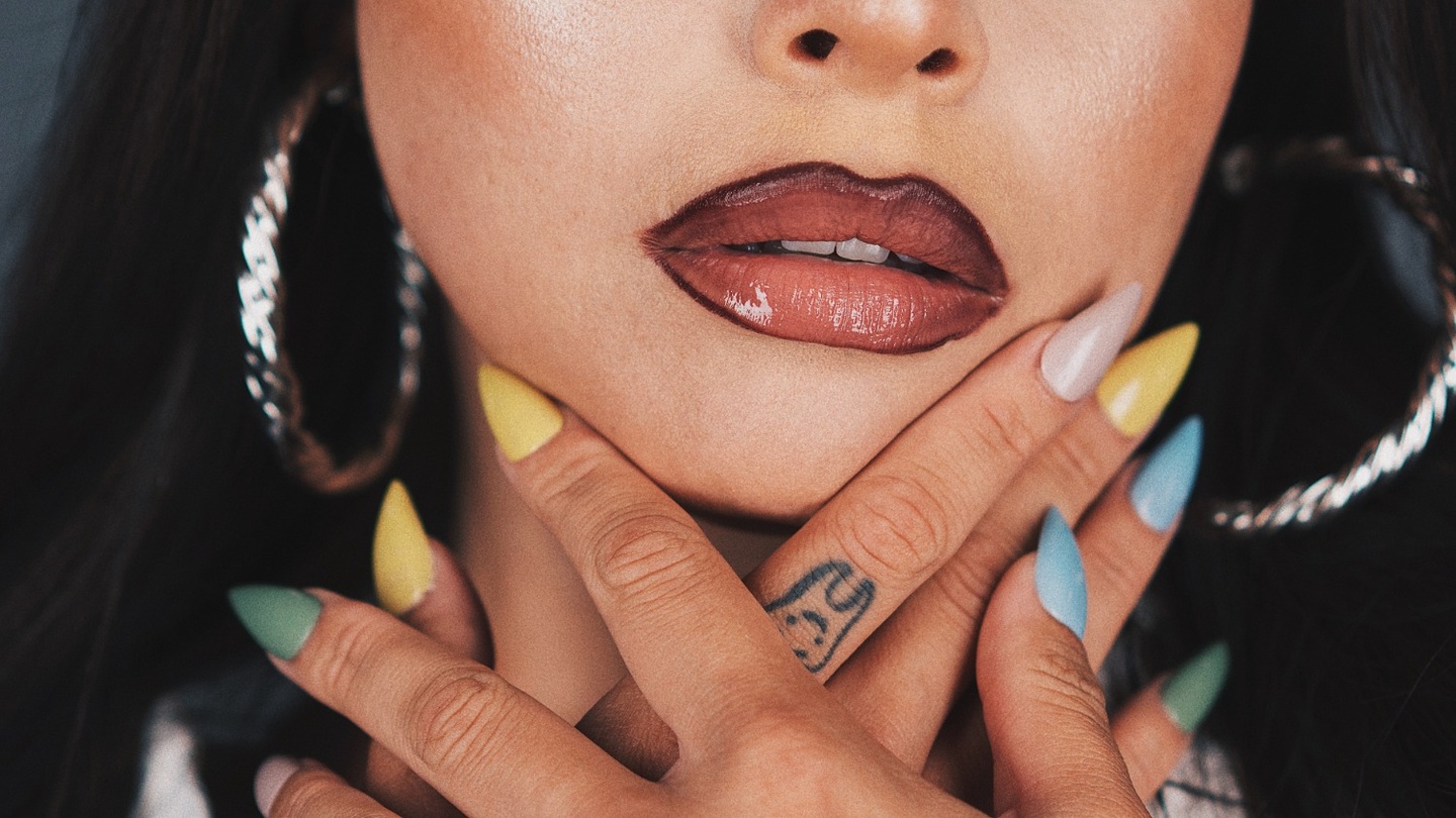 A close up of the bottom half of a face wearing dark lipstick with hands with pastel nails up by the chin.
