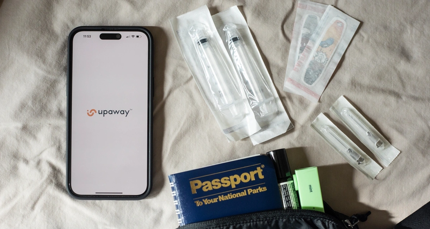A smartphone with the Upaway app open, passport, syringes, needles, and band aids are spread out on a bed sheet.