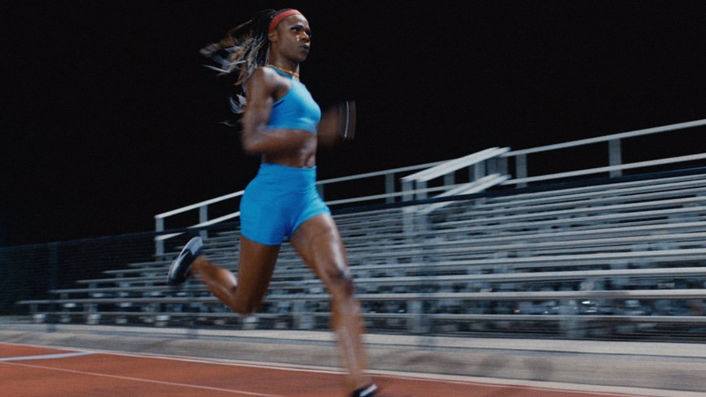 a black transgender track and field champion in a blue athletic gear running with bleachers behind her