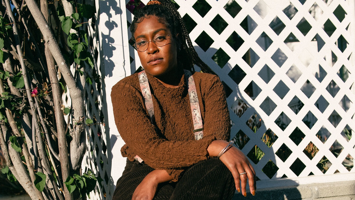 a black femme in a brown sweater staring at the camera in front of a latticework fence